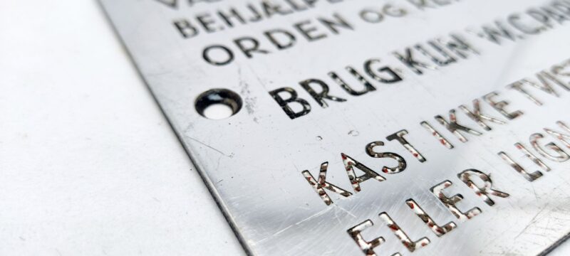 Metalskilt. Vær i egen interesse behjælpelig med at holde orden og renlighed. BRUG KUN W. C. PAPIR. Kast ikke tvist, klude eller lign. i kummen